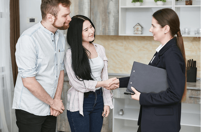 A real estate agent shakes hands with a smiling couple in a kitchen.
